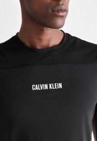 Man wearing a black Calvin Klein shirt with textured fabric across the shoulders and white logo text centered on the chest.