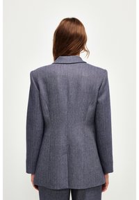 Blazer de arpillera de pata de gallo en tonos azul oscuro, con solapa muesca, hombros estructurados y un ajuste entallado con una textura suave.