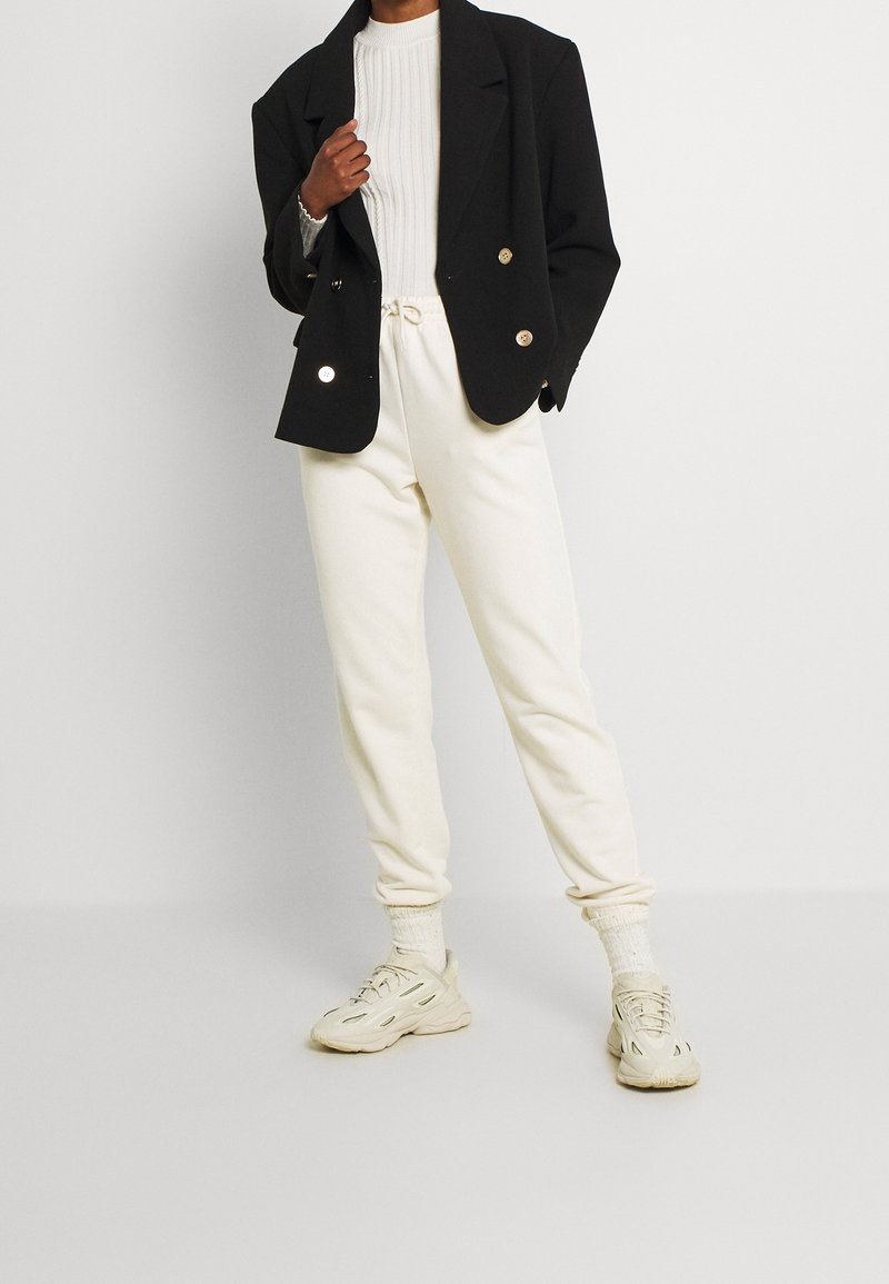 Personne portant un blazer noir croisé, un pull côtelé blanc, un pantalon de jogging crème, des chaussettes blanches et des baskets épaisses crème, debout sur un sol uni.