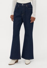 Flared dark blue denim jeans with front pockets, orange stitching, high waist, and raw hem, paired with white heeled boots.
