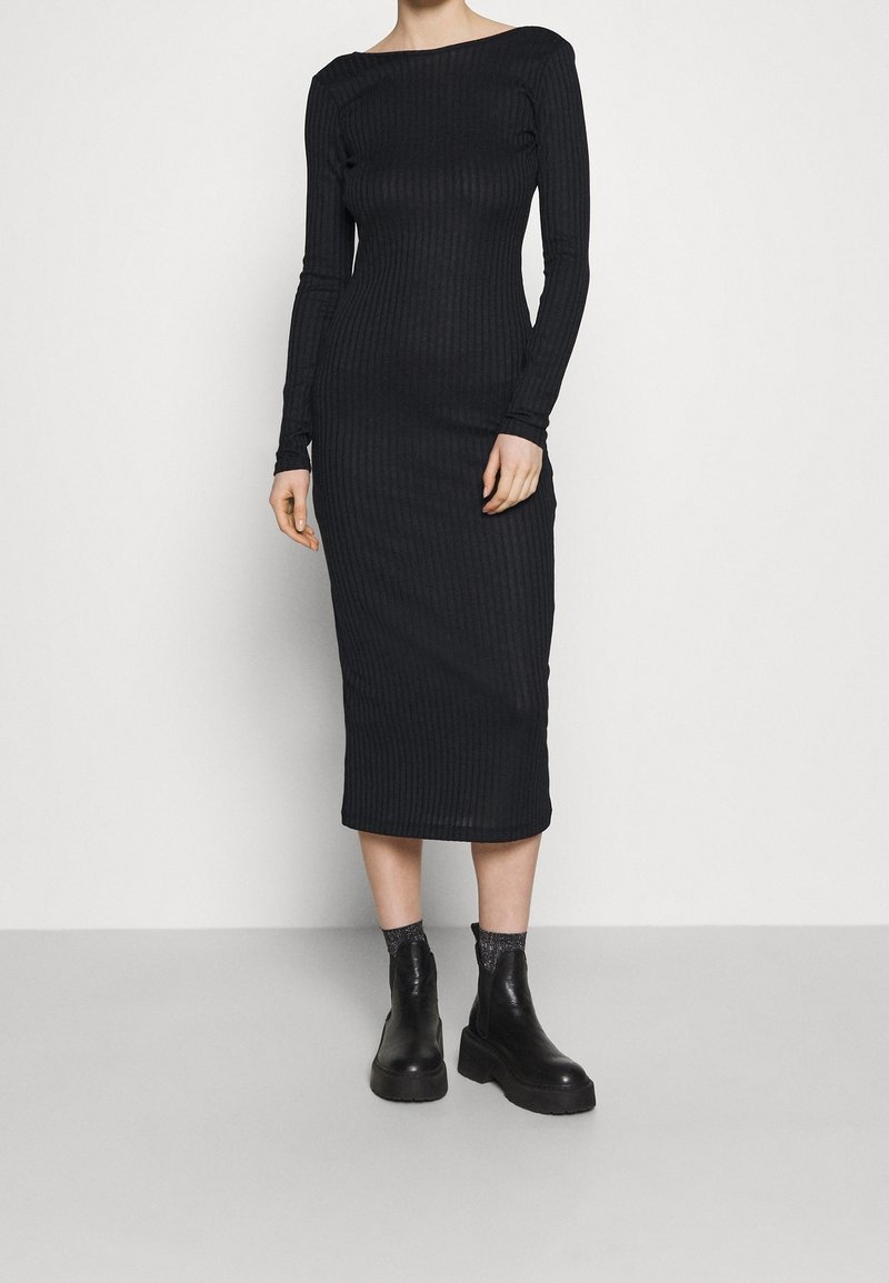 Femme portant une robe midi noire côtelée à manches longues, associée à des bottines noires et des chaussettes foncées, debout devant un fond clair uni.