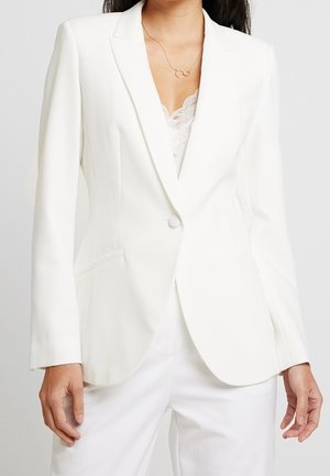 Vrouw met een op maat gemaakte witte blazer met één knoop, wit topje met kanten randje, witte broek en een delicate gouden ketting.