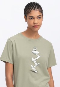 Lichtgroen katoenen t-shirt met een zwart grafisch ontwerp van gestapelde stenen en abstracte figuren. Ronde hals en korte mouwen.