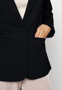 Personne portant un blazer noir avec une main dans la poche droite, un pantalon beige et un haut de couleur claire en dessous.