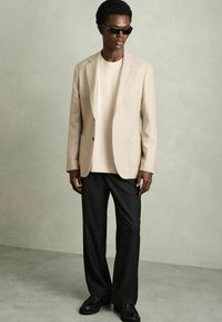 Blazer bej, croit pe un cămașă deschisă, asortat cu pantaloni negri cu croială largă și pantofi negri. Material neted cu un design structurat.