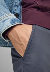 Chaqueta de denim de color azul claro con bordes deshilachados, superpuesta a un sudadera texturizada de color marrón oscuro, combinada con pantalones grises.