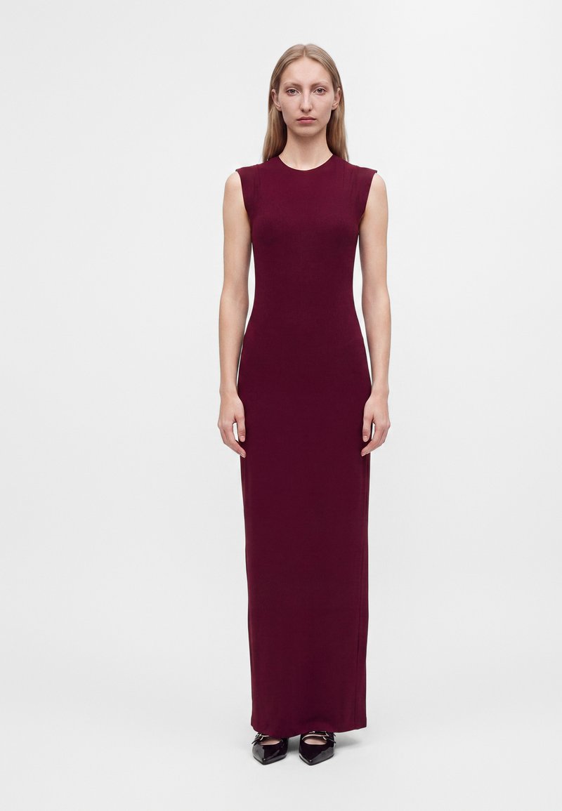 Femme debout, face à l'avant, portant une robe longue sans manches rouge foncé et des chaussures noires pointues sur un fond blanc uni.