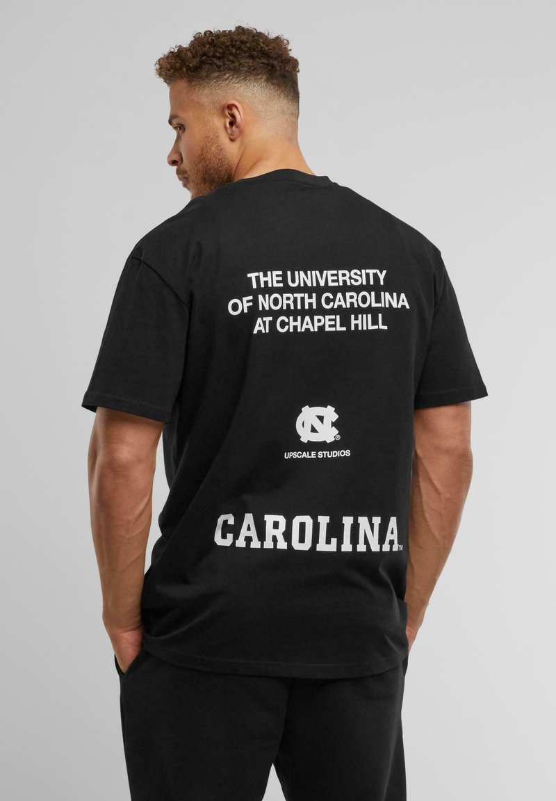 Musta puuvillainen t-paita, jonka selässä on valkoiset tekstit "NORTH CAROLINA YLIOPISTO CHAPEL HILLISSA" ja "CAROLINA."