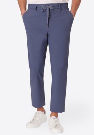 Uomo che indossa pantaloni blu su misura con vita con coulisse e sneakers bianche, in piedi con le mani in tasca contro uno sfondo bianco.
