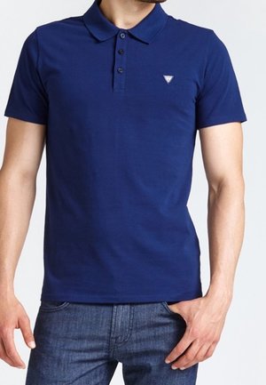 Homme portant un polo bleu marine ajusté à manches courtes, avec trois boutons et un petit logo triangulaire sur la poitrine.