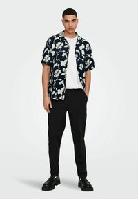 Kurzarmhemd mit navyfarbenem Hintergrund und weißen Blumenmustern, kombiniert mit schwarzen Hosen und schwarzen Schuhen, vor einem hellen Hintergrund stehend.
