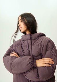 Puffige lila Jacke mit hohem Kragen und gerafften Ärmeln. Das Material wirkt weich und glatt und weist genähte Einsätze auf.