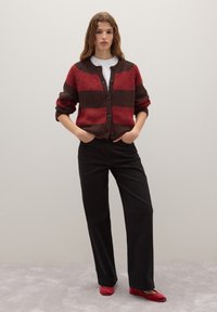 Cardigan à motifs rouges et bruns avec une texture côtelée, fermeture à boutons et manches retroussées, accompagné de pantalons larges noirs et de ballerines rouges.