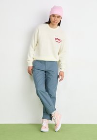 Kreemikäesed kampsun punase "DICKIES" logoga, sinised püksid, roosa beanie ja heledad roosad kingad valgete talladega, seistes rohelisel põrandal.