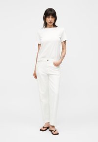 Femme aux cheveux foncés portant un t-shirt blanc simple, un pantalon blanc et des tongs noires, debout avec une main dans la poche.
