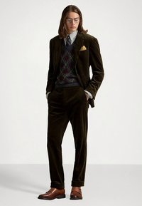 Κοστούμι από βελούδο σκούρο πράσινο με πουλόβερ μοτίβου και πουκάμισο με καρό, διακοσμημένο με κίτρινο μαντήλι στην τσέπη και καφέ παπούτσια.