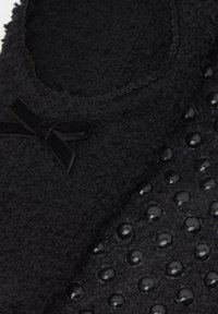 Pantofole nere con tessuto testurizzato, piccolo fiocco in velluto sopra e file di grip tattili rotondi sulla suola.