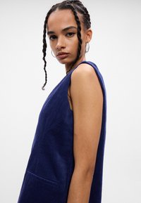 Bezrukávové šaty z tmavomodrého kordu s jemne rebrovým textúrou a bočnou kapsou, s vysokým výstrihom a voľným strihom.