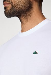 T-shirt bianco in cotone con colletto rotondo e logo di coccodrillo ricamato in verde sulla parte sinistra del petto. Tessuto liscio, vestibilità casual.
