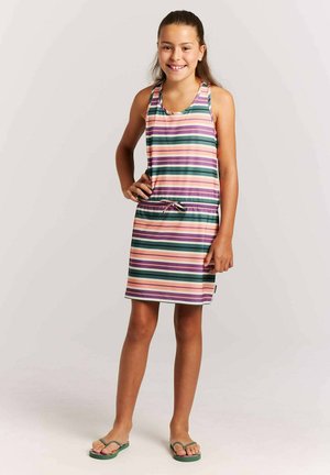 Niña de pie con la mano en la cadera, lleva un vestido sin mangas con rayas en púrpura, verde, rosa y beige, y chanclas verdes, sonriendo a la cámara.