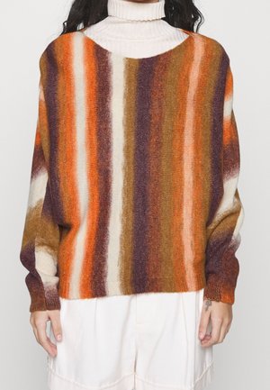Person iført en stribet, lodden sweater med lodrette bånd i orange, brun, lilla og creme over en hvid rullekrave og cremefarvede bukser.