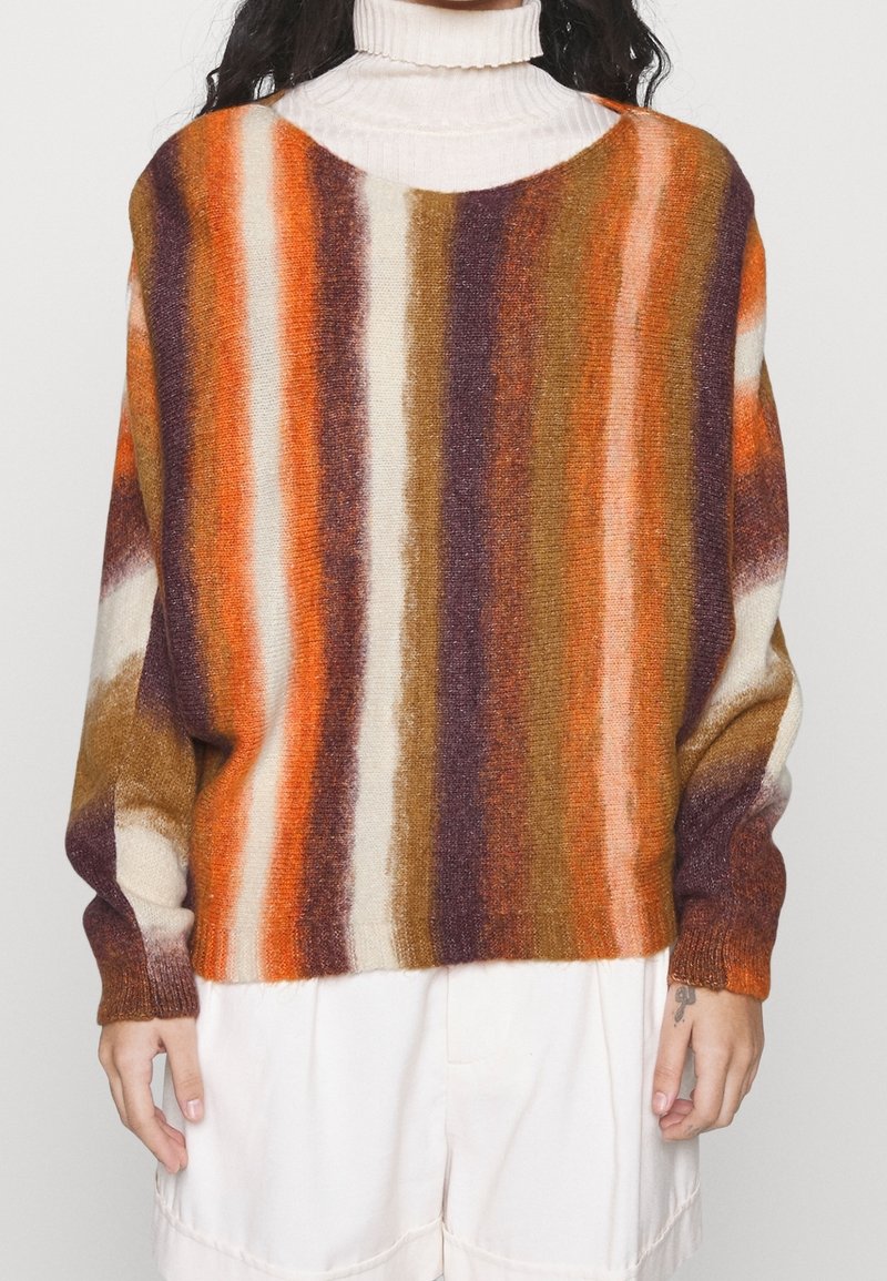 Personne portant un pull à rayures touffu avec des bandes verticales orange, marron, violet et crème, sur un col roulé blanc et un pantalon crème.