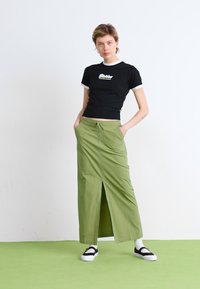 Čierne tričko s krátkym rukávom a bielym golierom, skombinované s dlhou olivovo zelenou sukňou s rozšírením a vreckami. Modelka má na sebe čierne sandále.