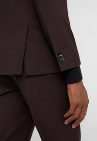 Donkerbruine colbert met een getailleerde pasvorm, voorzien van een split aan de achterkant, drie zwarte knopen op de mouw en een gebreide zwarte detail op de mouw.