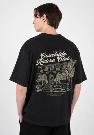 Ung mand set bagfra iført sort oversized t-shirt med "Courtside Riviera Club" og bygningstegning på ryggen.