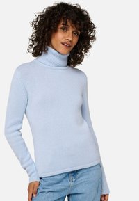 Femme aux cheveux bouclés portant un pull léger bleu avec un col montant et un jean bleu clair, posant avec une main sur la hanche.