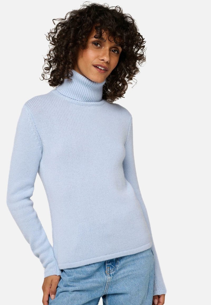 Femme aux cheveux bouclés portant un pull léger bleu avec un col montant et un jean bleu clair, posant avec une main sur la hanche.