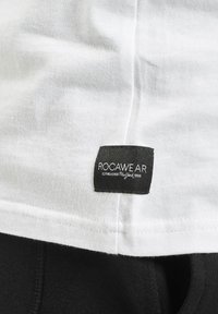 Un tricou alb cu o etichetă neagră vizibilă pe care scrie "ROCAWEAR" împreună cu "ESTABLISHED New York 1999." Materialul pare moale și ușor.