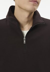 Mann in einem dunkelbraunen Viertelreißverschluss-Pullover über einem weißen gerippten Shirt, das seinen unteren Gesichtsteil und Hals vor einem weißen Hintergrund zeigt.