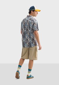 Krátky rukáv košeľa s modrým listovým vzorom, béžové cargo šortky, teal a biele tenisky, ponožky s viacfarebnými pruhmi a modro-žltá čiapka.