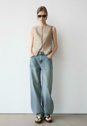 Frau trägt ärmellose beige Weste mit Knöpfen, weit geschnittene ausgewaschene blaue Jeans, schwarze Sonnenbrille und schwarze Zehensandalen, steht vor einer weißen Wand.