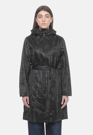 Donna con capelli castani lisci che indossa un cappotto nero lucido con cappuccio e cintura in vita, in piedi davanti a uno sfondo chiaro e semplice.