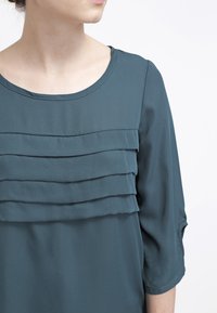 Blouse teal avec trois couches de volants horizontales sur la poitrine. Dotée d'un col rond et de manches 3/4. Texture lisse, tissu léger.