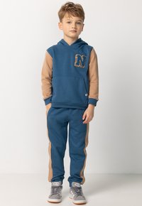 Blaues Kapuzensweatshirt mit braunen Ärmeln und einem "N"-Aufnäher, kombiniert mit passenden blauen Jogginghosen mit braunen seitlichen Streifen und Bündchen.