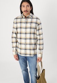 Camisa de franela a cuadros beige y azul marino con cuello abotonado, mangas largas y un pequeño logo en el pecho; combinada con jeans azules.