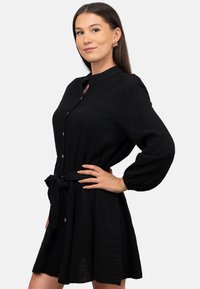 Abito nero realizzato in tessuto testurizzato con collettino, chiusura frontale con bottoni e maniche lunghe. Presenta una cintura da annodare in vita per un look aderente.