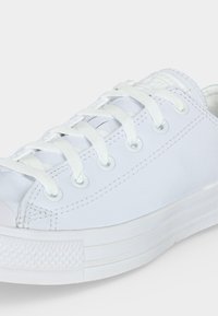 Λευκό low-top αθλητικό παπούτσι με λευκά κορδόνια, μεταλλικά ροκανίσματα, λαστιχένια σόλα και υφή στην άκρη, απεικονισμένο από κοντινή πλευρική γωνία.