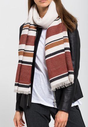 Femme portant une écharpe rayée marron, blanche, noire et beige sur une chemise blanche et une veste en cuir noire, avec un pantalon foncé.