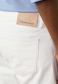 Jeans bianchi con una tasca posteriore visibile. Un'etichetta in pelle beige con la scritta "FACONNABLE" impressa in lettere scure è attaccata vicino alla vita.