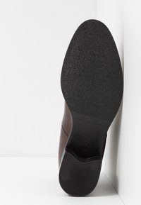 Scarpa in pelle marrone con suola in gomma testurizzata, punta arrotondata e design minimalista. Dettagli di cucitura lungo il lato sono visibili.