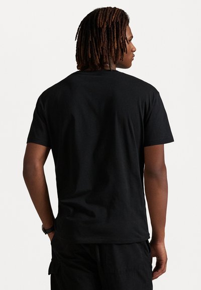 Man med axellång dreadlocks som bär en svart t-shirt och svarta byxor, vänd bort från kameran mot en enfärgad bakgrund.