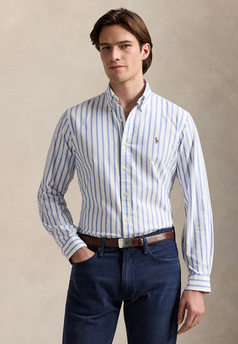 Bela srajca z ozkimi modrimi črtami, gumbi na ovratniku in izvezenim logotipom na prsih. Oblečena z temnimi denim jeans in rjavim pasom.