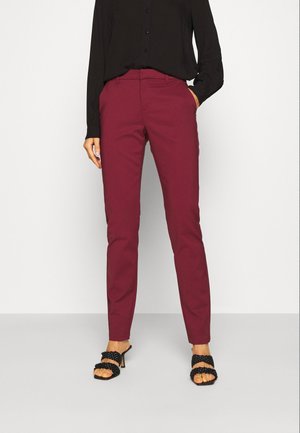 Vrouw draagt slanke bordeauxrode broek, zwart shirt met lange mouwen en zwarte sandalen met hak, staand tegen een effen witte achtergrond.
