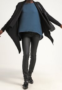 Černý kardigan s otevřeným předním dílem, modrozelený top, černé úzké kalhoty a černé boty. Hladké látky, volný střih, kontrastní barevné detaily.