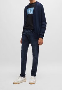 Marineblauer Cardigan mit Knöpfen, schwarzes T-Shirt mit blauem Grafikdruck, dunkle Jeans und weiße Sneakers mit hellbraunen Akzenten.
