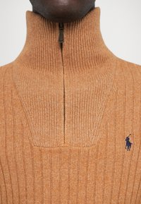 Pomarańczowy sweter z prążkowanej dzianiny, z wysokim kołnierzem i zamkiem błyskawicznym przy szyi. Posiada małe niebieskie logo na piersi. Miękka textura.